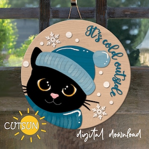 May include: A round wooden sign featuring a black cat wearing a blue hat and scarf. The sign reads "It's cold outside".