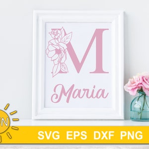 May include: A white frame with a pink floral design and the letter M, with the name Maria written below. The frame is sitting on a white surface.