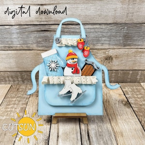 May include: A blue apron with a pocket filled with winter-themed decorations, including a snowman, mittens, ice skates, and a sled. The apron is on a wooden stand and the text "digital download" and "CUTSUN" are visible.