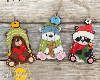 May include: Three wooden Christmas ornaments featuring cartoon animals. A brown bear with a green and yellow striped hat holds a red heart. A polar bear with a blue hat and green scarf holds a gingerbread man. A panda in a Santa hat holds a Christmas tree. The words "digital download" are at the top.