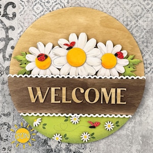 Peut inclure: Panneau de bienvenue rond en bois avec trois grandes marguerites blanches, des centres jaunes et des coccinelles rouges. Le mot "WELCOME" est affiché sur une bannière en bois marron. Une section verte en bas présente des accents de marguerites et de coccinelles.