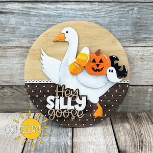 Könnte beinhalten: Runder Holzschild mit einer weißen Gans, die Süßigkeiten und einen Kürbis hält. Der Schriftzug "Hey Silly Goose" ist auf dem Schild zu sehen, ideal für Halloween-Dekorationen.
