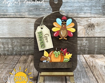 Può includere: Un tagliere in legno a tema del Ringraziamento con un tacchino, una zucca e decorazioni di foglie. La tavola ha un'etichetta con la scritta "Give Thanks". Le parole "digital download" sono scritte in alto. La tavola è su un cavalletto di legno.