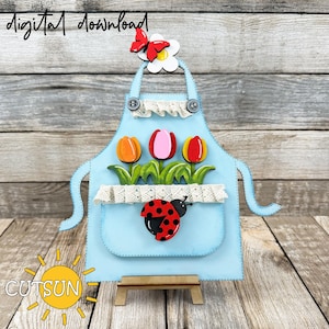 May include: A blue wooden apron with a white lace trim and a pocket. The apron has a ladybug, three tulips, and a butterfly with a white flower on top. The apron is on a small wooden stand.