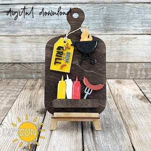 May include: A wooden cutting board with a brown background and a yellow tag that says "King of the Grill". There are also images of a grill, flames, mustard, ketchup, mayonnaise, and a sausage on the board. The board is on a small wooden easel.