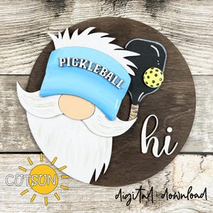 May include: A wooden round sign featuring a gnome with a white beard and a blue hat that says "PICKLEBALL". A black pickleball paddle and yellow ball are on the right. The word "hi" is written in white.