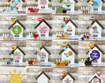 May include: A collection of 12 wooden house-shaped signs with various seasonal and holiday themes. Each sign features a different design, including a snowman, a heart, a rainbow, a bunny, a school bus, a scarecrow, a ghost, a pumpkin, a turkey, and a Christmas tree. The signs are all painted in bright colours and have the word "HOME" written on them.
