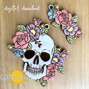 May include: A white skull with a black outline is decorated with pink, yellow, and purple flowers. The skull has a black eye socket and a black mouth with teeth. The flowers are arranged around the skull and are in a floral pattern. The skull is on a wooden background. "Digital download" is written in white text on the top left of the image.