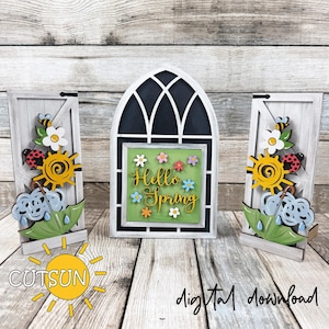 May include: Wooden spring-themed decorations. A central arched window with "Hello Spring" text is flanked by two door-shaped panels with sun, bee, ladybug, and umbrella designs. The colour palette includes white, green, yellow, and blue.