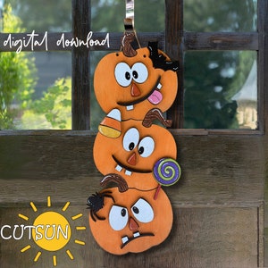 Puede incluir: Una pila de tres calabazas naranjas con caras tontas, cada una con diferentes detalles. La calabaza de arriba tiene un murciélago, la calabaza del medio tiene un maíz dulce y la calabaza de abajo tiene una araña y una piruleta.