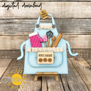 May include: A blue apron with a white lace trim and a pocket that says "Mom's Kitchen". The apron is decorated with a pink oven mitt, wooden kitchen utensils, and a wooden sign that says "The Kitchen - My Rules".