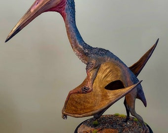May include: A detailed sculpture of a Pterosaur, painted in shades of brown, gold, blue, and red. The artwork is mounted on a round base with small rocks and grass. The text "Painted by: Jarrod Harvey" is visible.