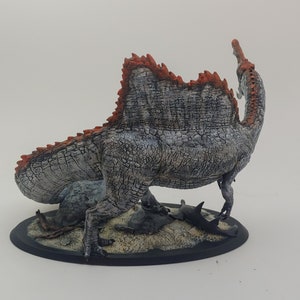 Puede incluir: Una figurita de dinosaurio pintada de gris y naranja con una espalda espinosa, de pie sobre una base negra con una textura arenosa. El dinosaurio est&aacute; representado con un cuello y una cola largos, y su boca est&aacute; abierta.