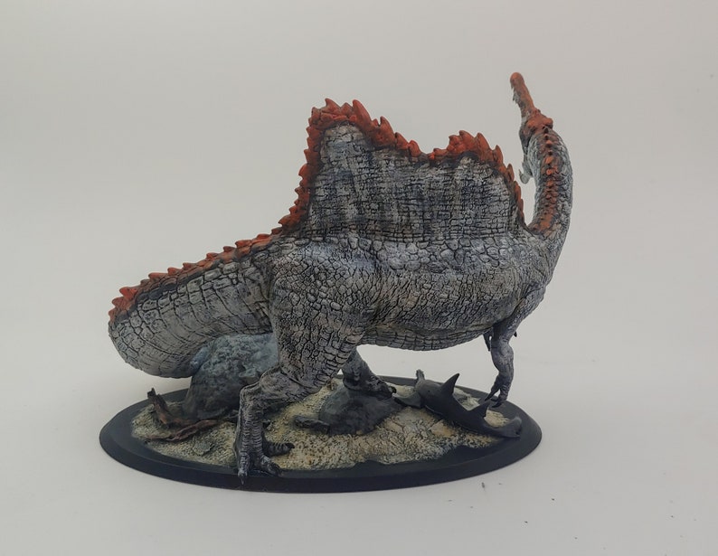 Puede incluir: Una figurita de dinosaurio pintada de gris y naranja con una espalda espinosa, de pie sobre una base negra con una textura arenosa. El dinosaurio est&aacute; representado con un cuello y una cola largos, y su boca est&aacute; abierta.