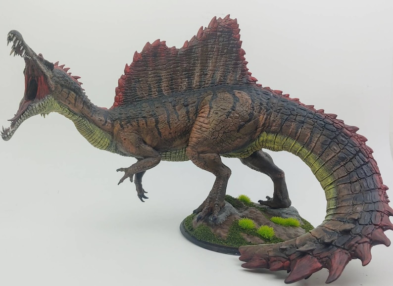 Puede incluir: Una miniatura detallada y pintada a mano de un dinosaurio Spinosaurus. El dinosaurio est&aacute; representado en una pose din&aacute;mica con la boca abierta, revelando dientes afilados. La figura est&aacute; pintada en tonos de marr&oacute;n, verde y rojo, con una vela prominente en su espalda.