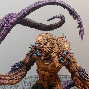 May include: A two-headed, brown and gold monster with a purple and brown tentacle-like arm. The monster has sharp teeth and blue eyes.