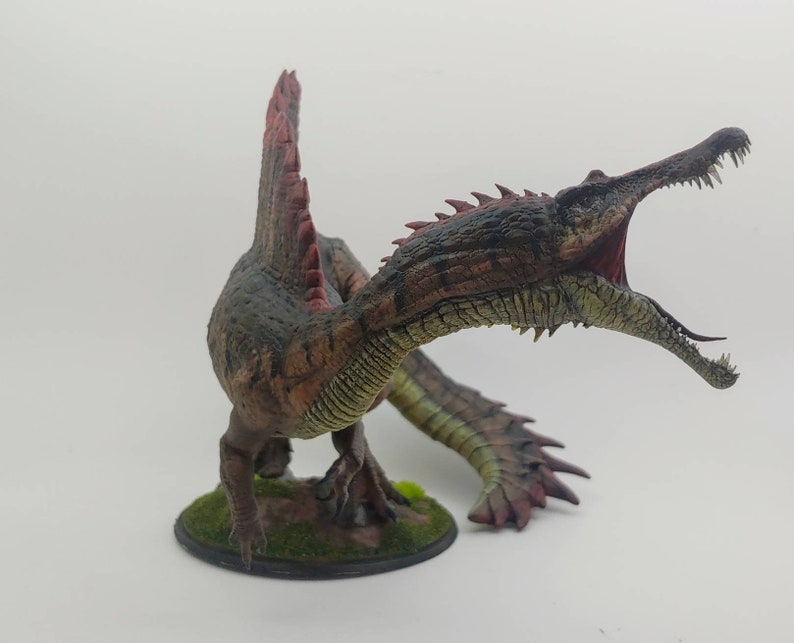 Puede incluir: Una miniatura detallada y pintada a mano de un dinosaurio Spinosaurus con un hocico largo y estrecho, una vela en la espalda y una cola larga y delgada. El dinosaurio est&aacute; pintado en tonos de marr&oacute;n, verde y rojo, y est&aacute; posado con la boca abierta en un rugido.