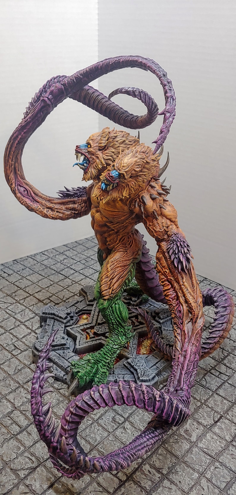 May include: A multi-headed, multi-limbed creature with a lion-like head and a green, scaly tail. The creature is painted in shades of brown, orange, and purple, and is standing on a stone base.