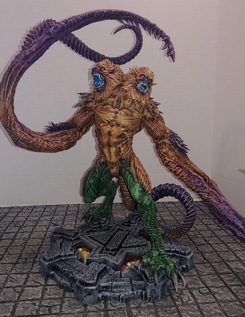 May include: A two-headed, multi-limbed creature with a yellow, green, and purple paint scheme. The creature is standing on a gray, stone base with a textured surface.