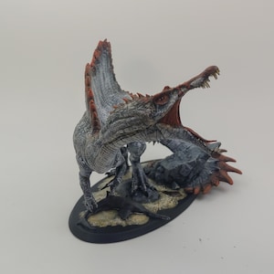 Puede incluir: Una miniatura pintada de un dinosaurio Spinosaurus con un cuerpo blanco y gris, aletas rojas y naranjas, y una boca abierta con dientes afilados. El dinosaurio est&aacute; de pie sobre una base negra con textura de roca gris y arena.