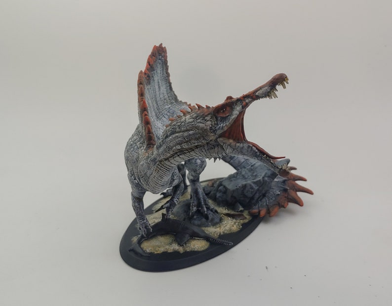 Puede incluir: Una miniatura pintada de un dinosaurio Spinosaurus con un cuerpo blanco y gris, aletas rojas y naranjas, y una boca abierta con dientes afilados. El dinosaurio est&aacute; de pie sobre una base negra con textura de roca gris y arena.
