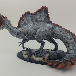 Puede incluir: Una miniatura pintada a mano de un dinosaurio Spinosaurus con un cuerpo gris y blanco, espinas rojas y naranjas y una cola larga. El dinosaurio est&aacute; en posici&oacute;n de rugido, con la boca abierta y la cabeza echada hacia atr&aacute;s. La miniatura est&aacute; colocada sobre una base con una peque&ntilde;a roca gris y una textura arenosa.