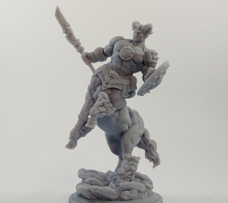 Puede incluir: Una miniatura gris de un guerrero centauro impresa en 3D, sosteniendo una lanza y un escudo. El centauro est&aacute; de pie sobre una base rocosa.