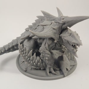 Pode incluir: Uma miniatura cinzenta de uma criatura fantástica impressa em 3D com uma concha espinhosa e uma cauda longa. A criatura está segurando uma criatura menor em suas garras.