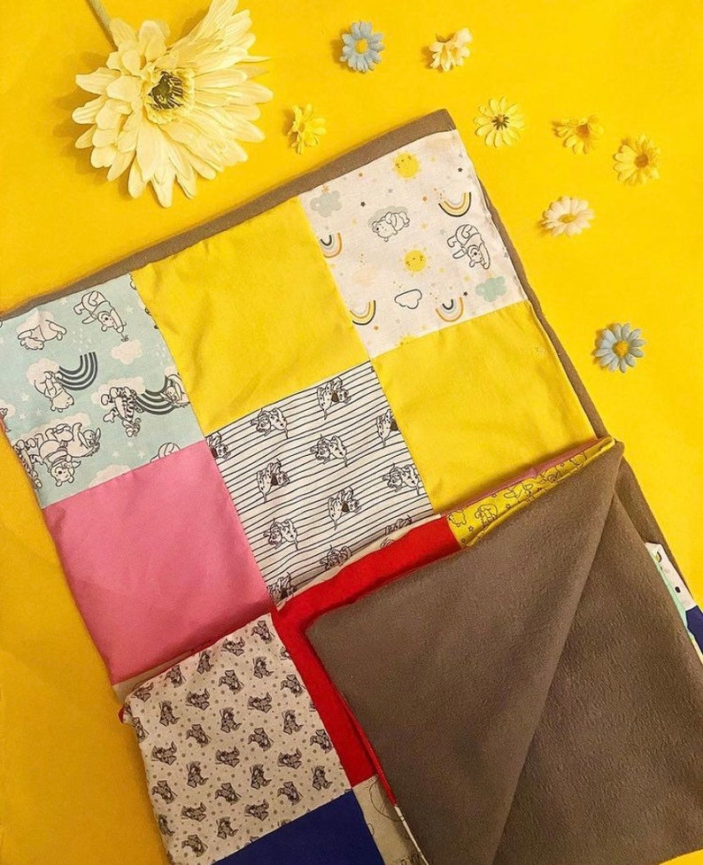 Peut inclure: Une couverture patchwork pour b&eacute;b&eacute; avec des carr&eacute;s de tissu jaunes, roses, bleus et &agrave; motifs. La couverture a un envers en polaire grise et est d&eacute;cor&eacute;e de petites fleurs.