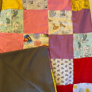 Peut inclure: Une courtepointe patchwork avec divers carr&eacute;s de tissu jaunes, roses, rouges et &agrave; motifs. Les motifs comprennent des personnages de dessins anim&eacute;s, des timbres et des illustrations de chiens. Une couverture en polaire grise recouvre partiellement la courtepointe.