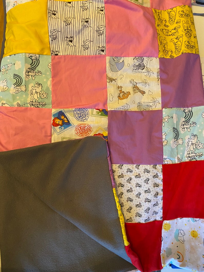 Peut inclure: Une courtepointe patchwork avec divers carr&eacute;s de tissu jaunes, roses, rouges et &agrave; motifs. Les motifs comprennent des personnages de dessins anim&eacute;s, des timbres et des illustrations de chiens. Une couverture en polaire grise recouvre partiellement la courtepointe.