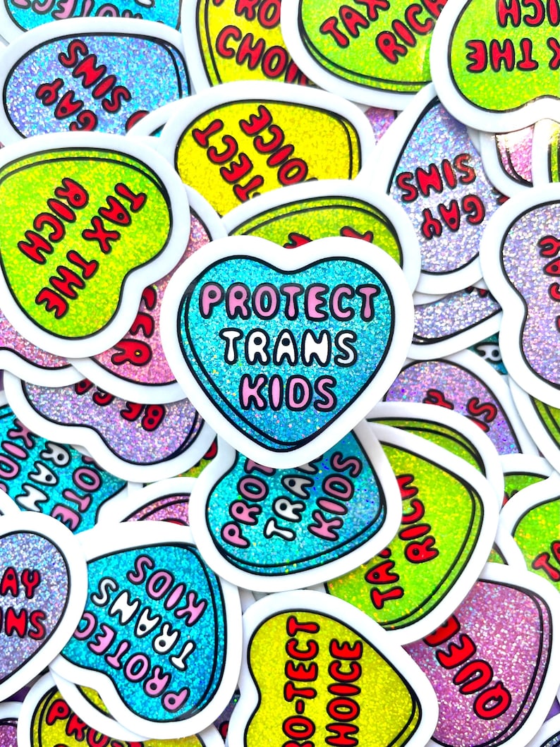 Pode incluir: Uma cole&ccedil;&atilde;o de autocolantes em forma de cora&ccedil;&atilde;o com fundos brilhantes e bordas brancas. Os autocolantes apresentam frases como "Protect Trans Kids", "Tax the Rich" e "Gay Sims". Os autocolantes s&atilde;o em v&aacute;rias cores, incluindo azul, verde, amarelo e rosa.