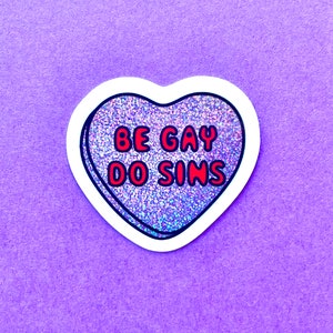 Pode incluir: Um autocolante em forma de cora&ccedil;&atilde;o com o texto "BE GAY DO SINS" em letras vermelhas com contorno preto. O cora&ccedil;&atilde;o tem um preenchimento brilhante e iridescente e uma borda branca. O autocolante est&aacute; num fundo roxo.
