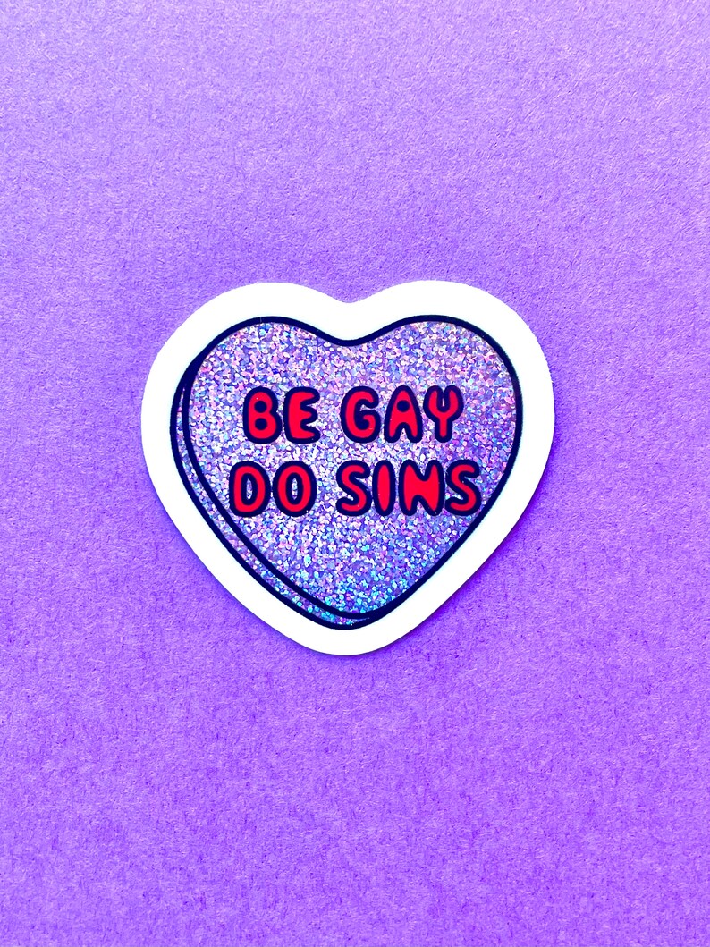 Pode incluir: Um autocolante em forma de cora&ccedil;&atilde;o com o texto "BE GAY DO SINS" em letras vermelhas com contorno preto. O cora&ccedil;&atilde;o tem um preenchimento brilhante e iridescente e uma borda branca. O autocolante est&aacute; num fundo roxo.
