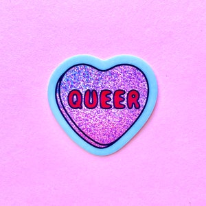 Pode incluir: Um autocolante em forma de cora&ccedil;&atilde;o com uma borda azul clara e a palavra "QUEER" em letras vermelhas. O interior do cora&ccedil;&atilde;o &eacute; preenchido com material rosa e roxo brilhante e brilhante. O autocolante est&aacute; sobre um fundo rosa liso.