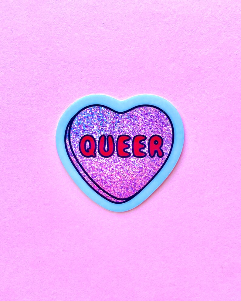 Pode incluir: Um autocolante em forma de cora&ccedil;&atilde;o com uma borda azul clara e a palavra "QUEER" em letras vermelhas. O interior do cora&ccedil;&atilde;o &eacute; preenchido com material rosa e roxo brilhante e brilhante. O autocolante est&aacute; sobre um fundo rosa liso.
