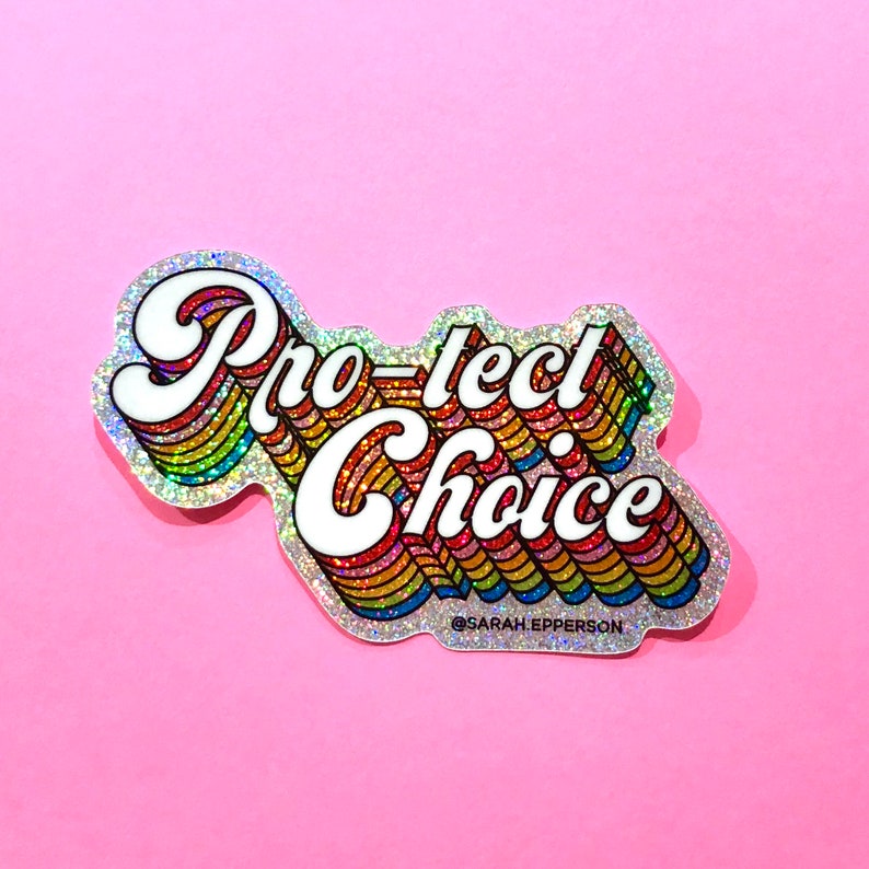 Pu&ograve; includere: Un adesivo glitter arcobaleno con il testo "Pro-tect Choice" in lettere bianche. L'adesivo ha un font retr&ograve; anni '70 ed &egrave; su uno sfondo rosa.