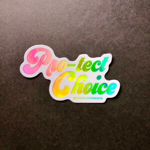 Pu&ograve; includere: Un adesivo bianco con finitura olografica che presenta il testo "Protect Choice" in un gradiente arcobaleno. Il testo &egrave; in uno stile di font retr&ograve;. L'adesivo include anche il testo "@SARAH_EPPERSON" in un font pi&ugrave; piccolo.