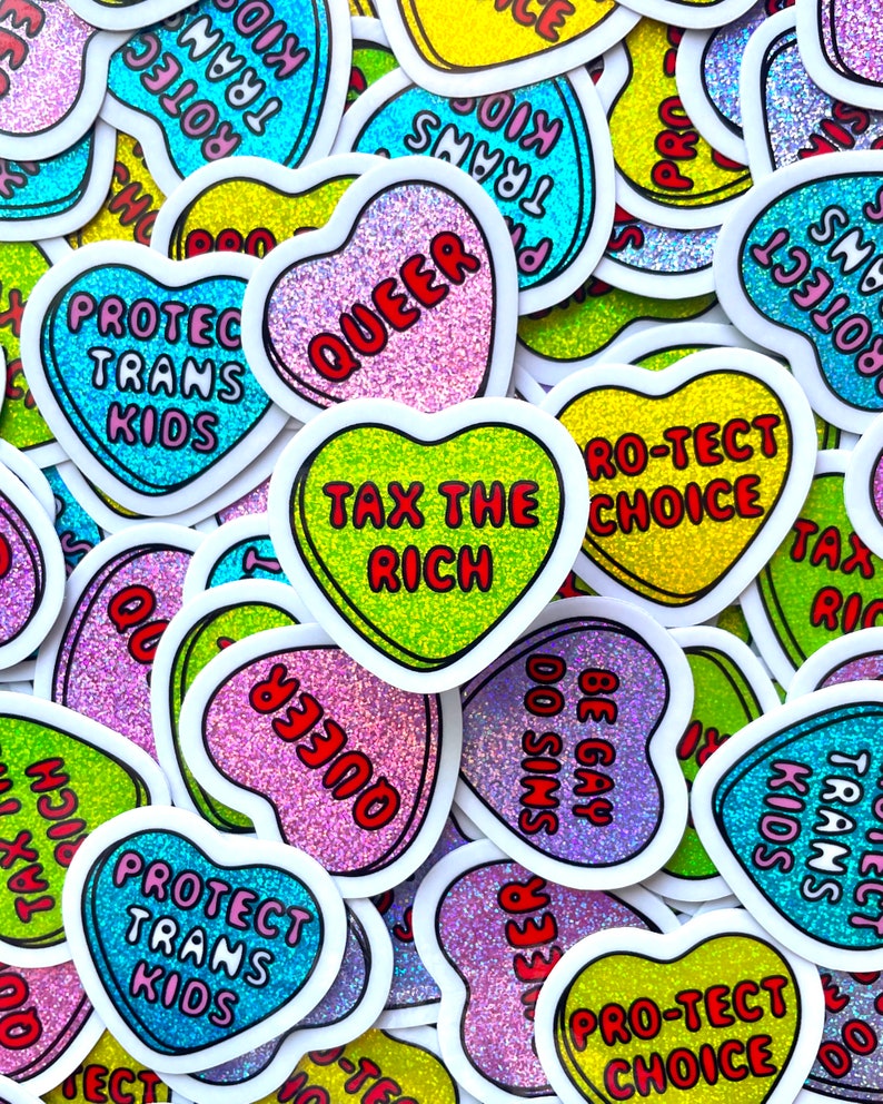 Pode incluir: Uma cole&ccedil;&atilde;o de autocolantes em forma de cora&ccedil;&atilde;o com fundos brilhantes e bordas brancas. Os autocolantes apresentam frases como "Protect Trans Kids", "Queer", "Tax the Rich" e "Be Gay Do Sins" em texto colorido.