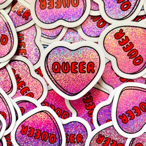 Pode incluir: Uma cole&ccedil;&atilde;o de autocolantes em forma de cora&ccedil;&atilde;o com um fundo rosa brilhante. Cada autocolante tem uma borda branca e a palavra "QUEER" em letras vermelhas. Os autocolantes est&atilde;o espalhados pela imagem.