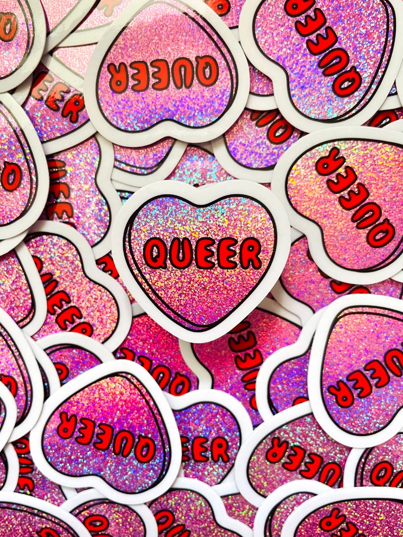 Pode incluir: Uma cole&ccedil;&atilde;o de autocolantes em forma de cora&ccedil;&atilde;o com um fundo rosa brilhante. Cada autocolante tem uma borda branca e a palavra "QUEER" em letras vermelhas. Os autocolantes est&atilde;o espalhados pela imagem.