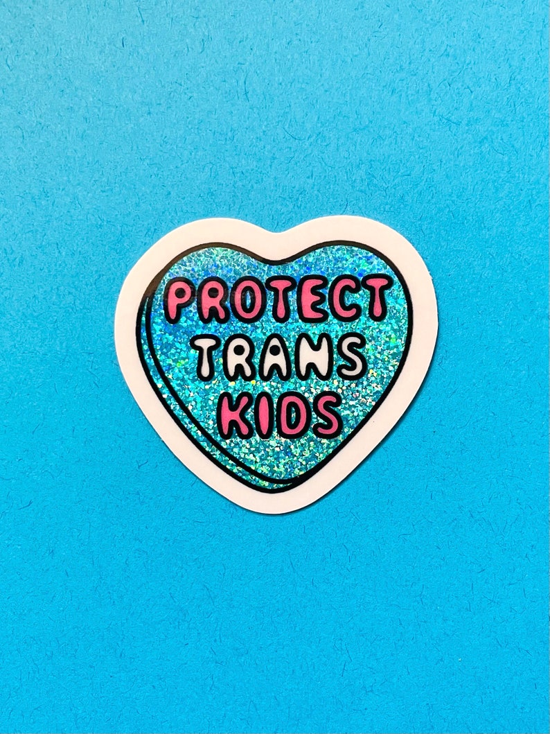 Pode incluir: Um autocolante em forma de cora&ccedil;&atilde;o com uma borda branca e um interior azul brilhante. O autocolante tem as palavras "PROTECT TRANS KIDS" em letras cor-de-rosa.