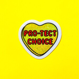 Pode incluir: Um autocolante em forma de cora&ccedil;&atilde;o com as palavras "PRO-TECT CHOICE" em letras vermelhas. O cora&ccedil;&atilde;o tem um fundo dourado brilhante e uma borda branca, sobre um fundo amarelo brilhante.