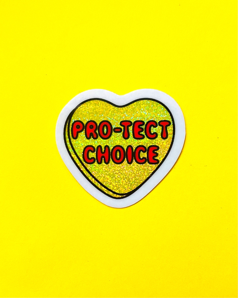 Pode incluir: Um autocolante em forma de cora&ccedil;&atilde;o com as palavras "PRO-TECT CHOICE" em letras vermelhas. O cora&ccedil;&atilde;o tem um fundo dourado brilhante e uma borda branca, sobre um fundo amarelo brilhante.