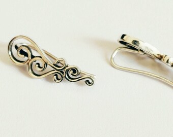 May include: A pair of silver ear cuffs with a delicate, swirling design. The cuff on the left has a more intricate design with multiple swirls, while the cuff on the right is a simple, curved shape.