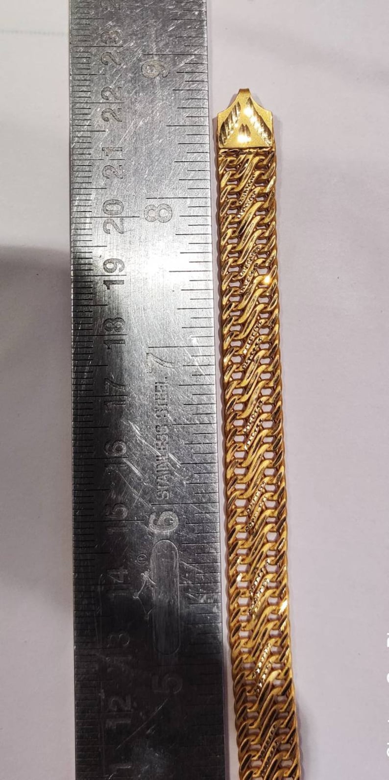 May include: A gold chain bracelet with a detailed, intricate design. The chain is measured against a ruler, showing its length.