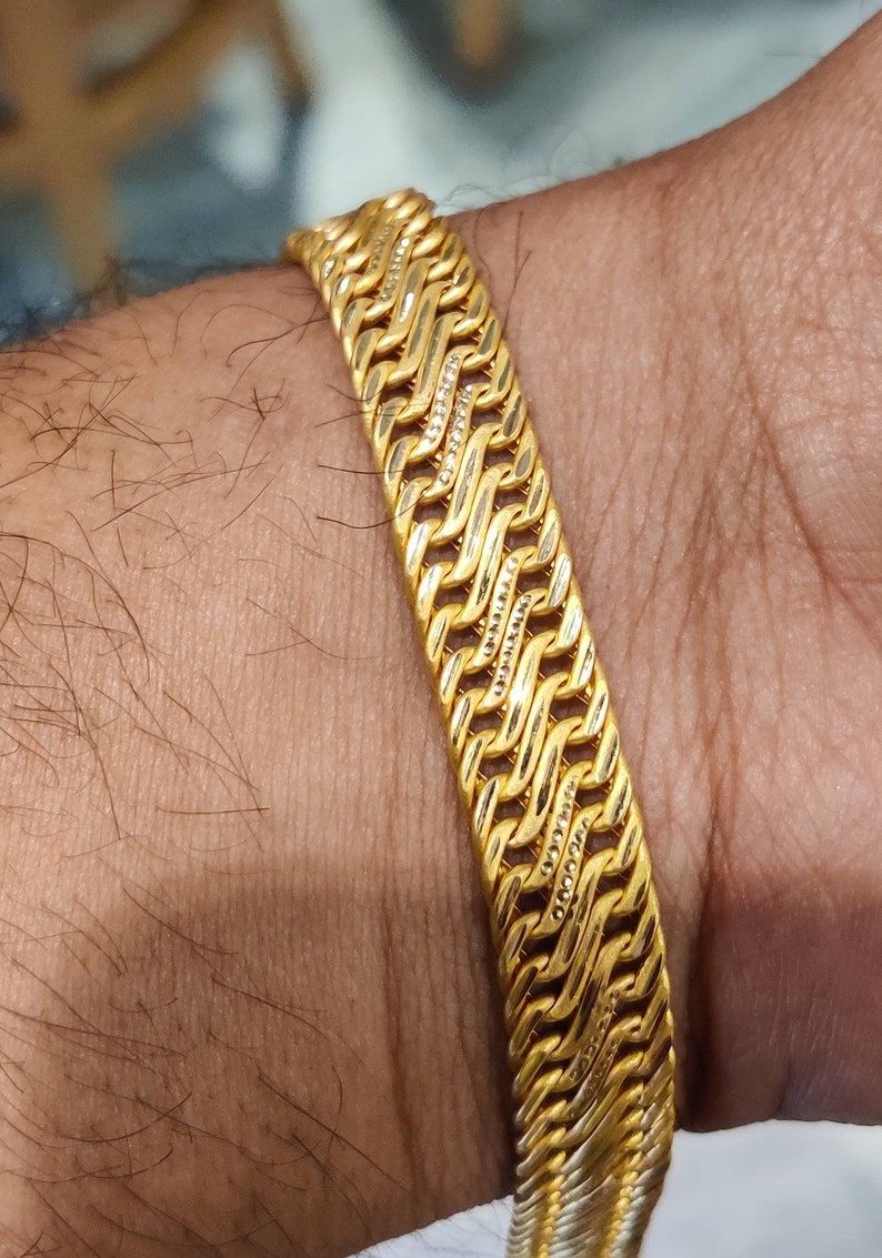 May include: A gold bracelet with a braided design. The bracelet is made of multiple strands of gold that are woven together to create a complex pattern.