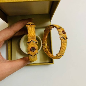 May include: Two gold bangles with intricate designs and red and green gemstones. The bangles are in a box with a white interior.