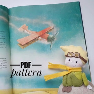 Op de afbeelding: Een vilten pop van een personage uit een kinderboek, met een gele hoed met een bril, een gele sjaal en groene kleding, zit op een pagina van een boek. Een vintage vliegtuigillustratie staat op de achtergrond. De tekst "PDF pattern" wordt weergegeven.