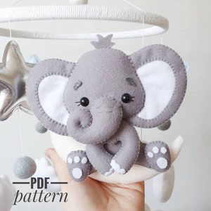Puede incluir: Un elefante de fieltro gris con orejas blancas y un corazón blanco en el pecho está sentado en una luna creciente de fieltro blanco. El elefante forma parte de un móvil con una estrella plateada y pompones grises. El texto "PDF pattern" está escrito debajo de la imagen.